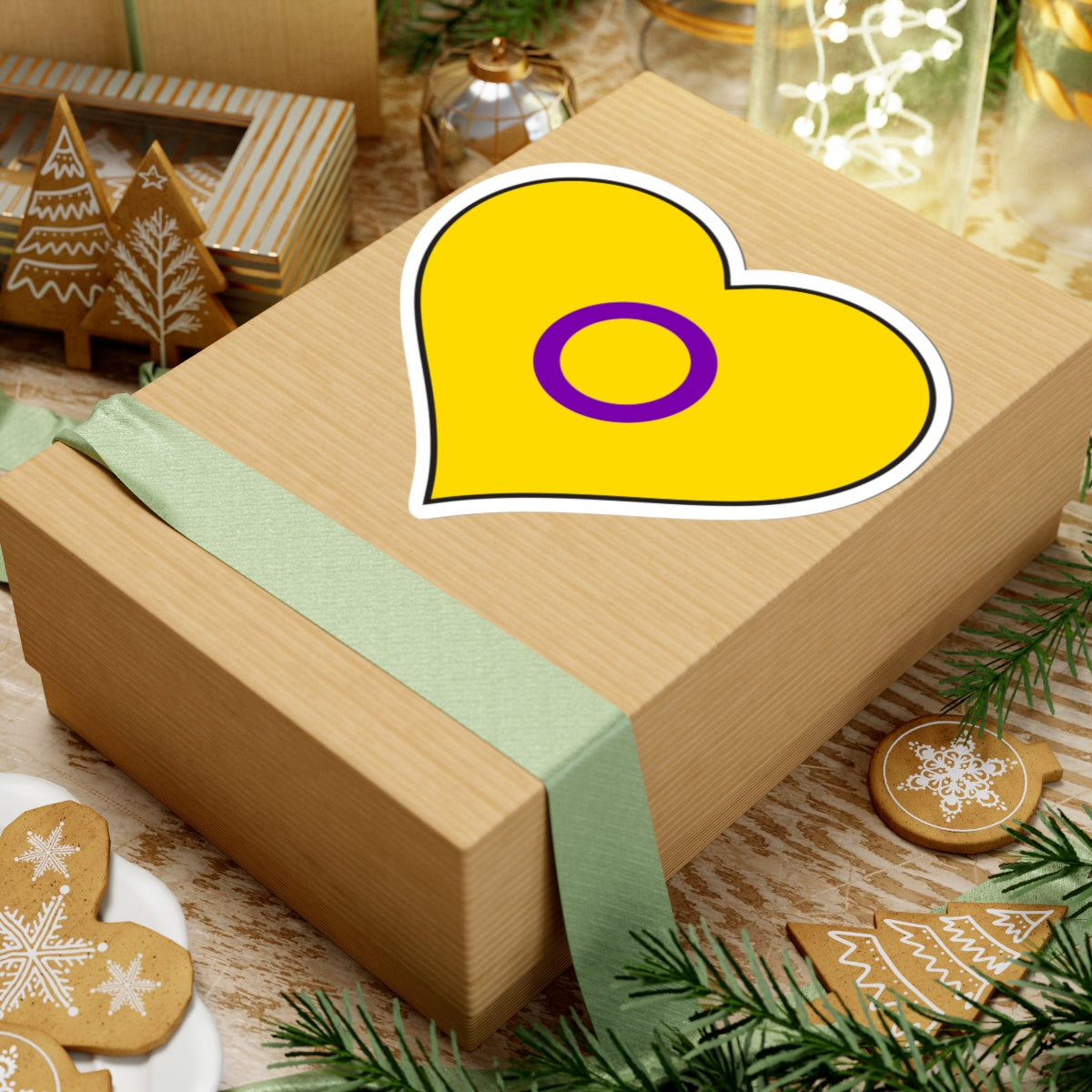 Gift box with a green ribbon on a festive background with cookies and Christmas decorations. Heart-shaped Intersex pride sticker centered on the box.