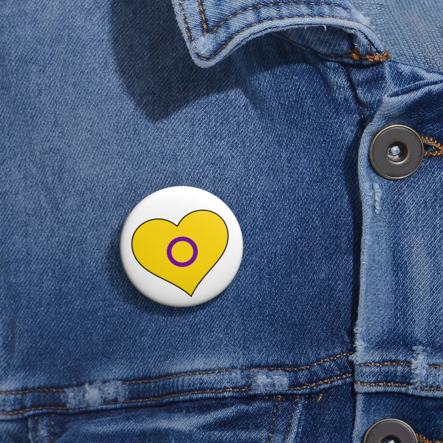 Close-up of a white circular pride button featuring a nested heart design inspired by the Intersex pride flag in yellow and purple, attached to a blue denim jacket at the right chest with the button clearly visible.