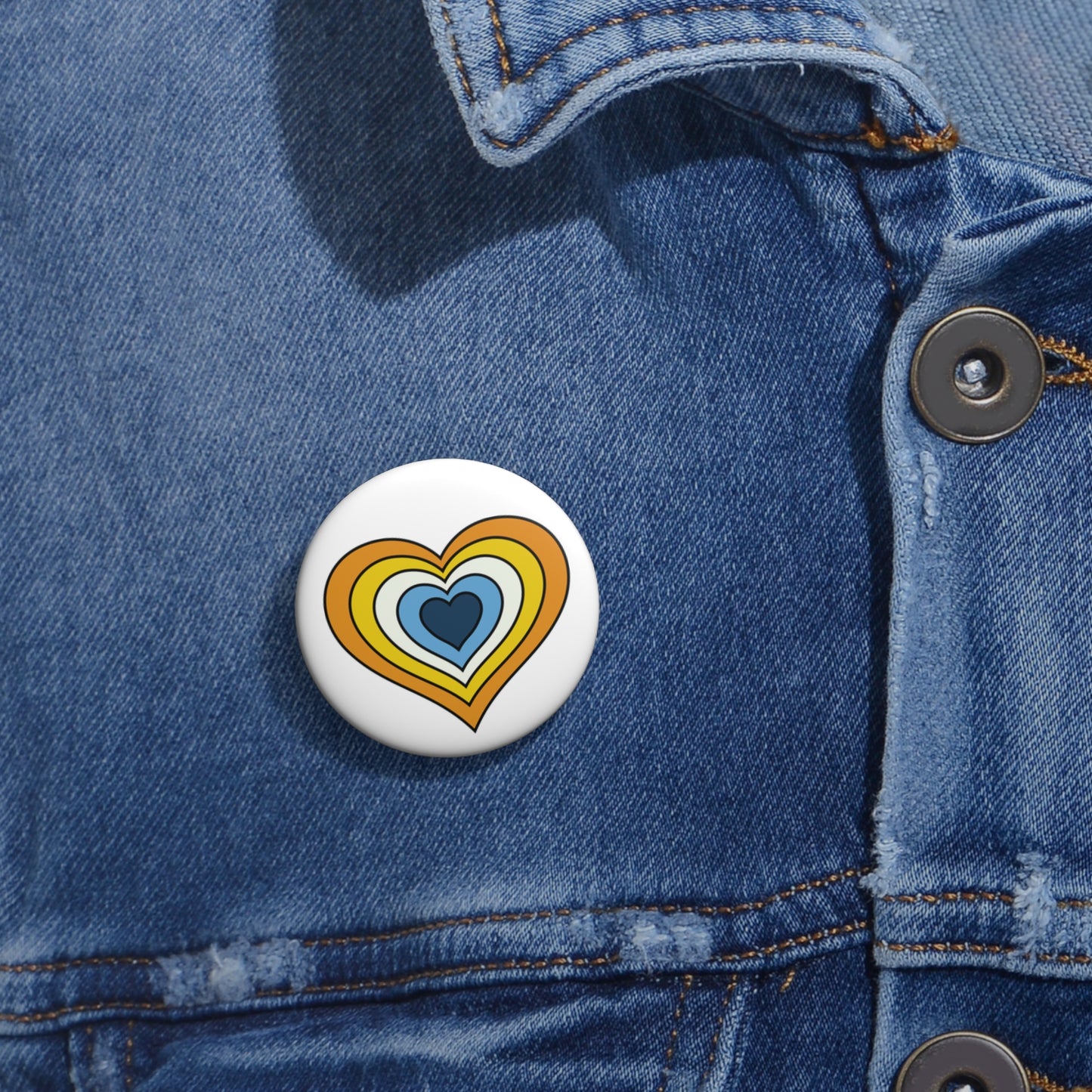 Close-up of a white circular pride button featuring a nested heart design inspired by the Aroace pride flag in yellows, white and blues, attached to a blue denim jacket at the right chest with the button clearly visible.