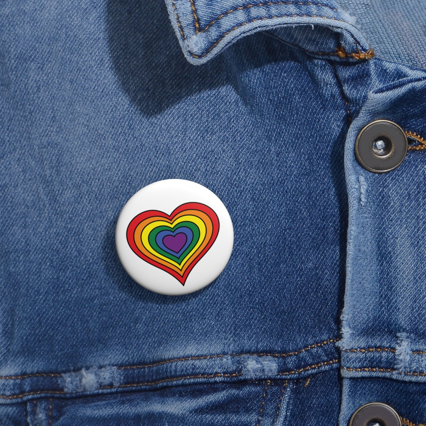 Close-up of a white circular pride button featuring a nested heart design inspired by the rainbow pride flag, attached to a blue denim jacket at the right chest with the button clearly visible.