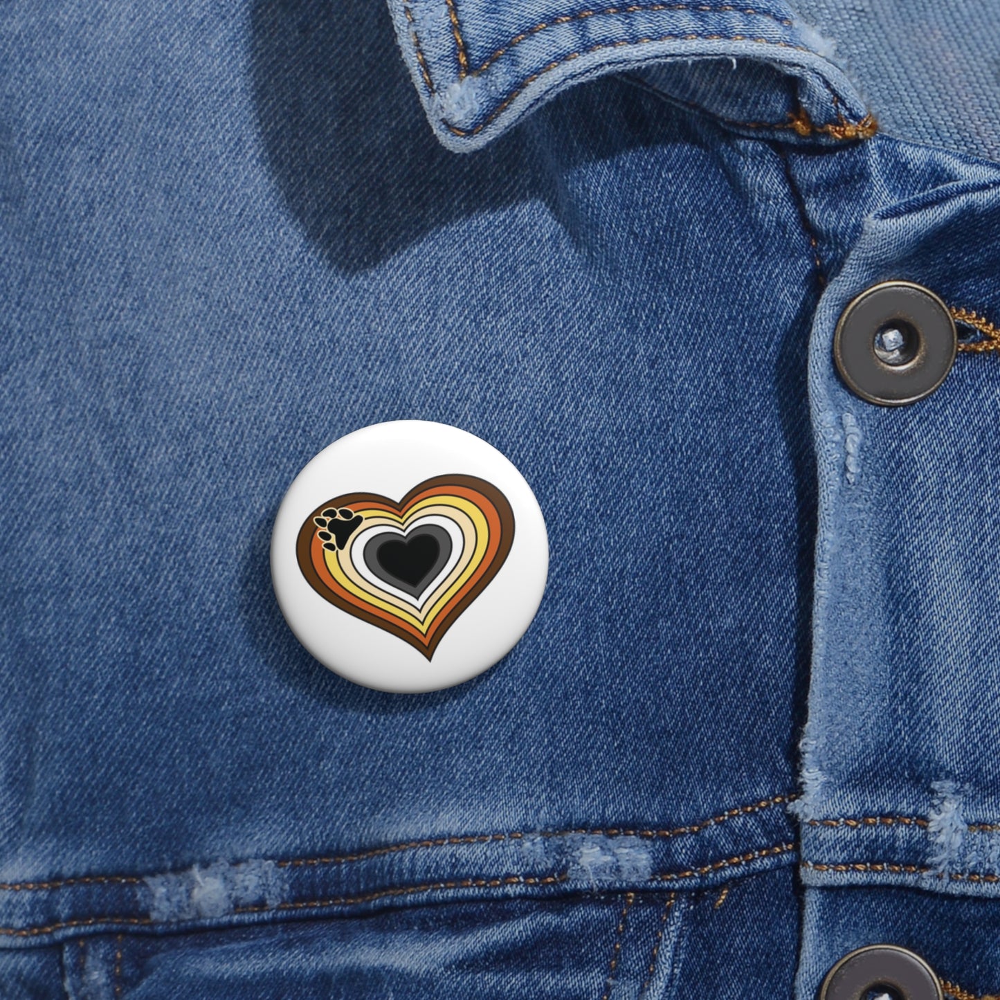 Close-up of a white circular pride button featuring a nested heart design inspired by the Bear pride flag in browns, cream, white, grey and black, attached to a blue denim jacket at the right chest with the button clearly visible.