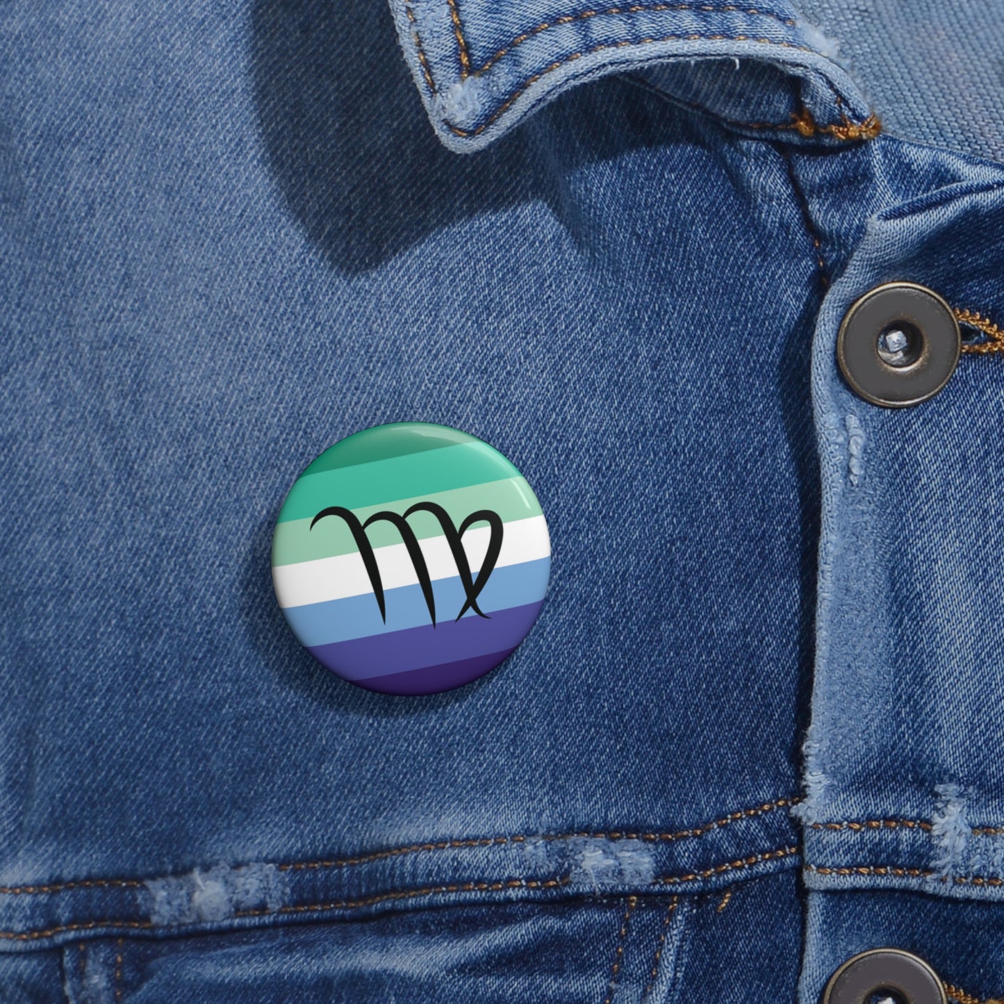 Close-up of a white circular pride button featuring the Virgo zodiac glyph centered over a gay men (MLM) pride flag background in greens/teals, white, and blues, attached to a blue denim jacket at the right chest with the button clearly visible.