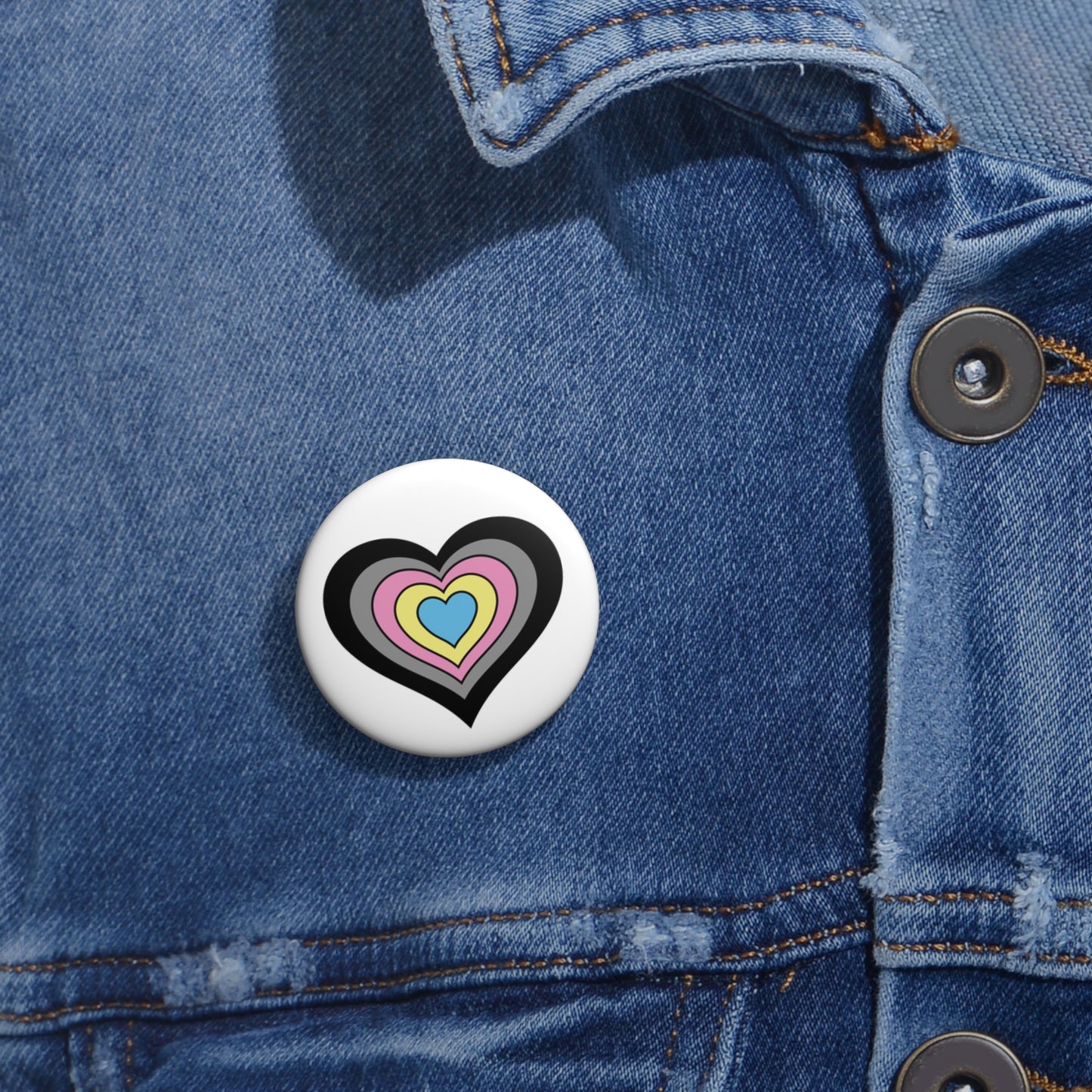 Close-up of a white circular pride button featuring a nested heart design inspired by the Polygender pride flag in black, gray, pink, yellow and blue, attached to a blue denim jacket at the right chest with the button clearly visible.