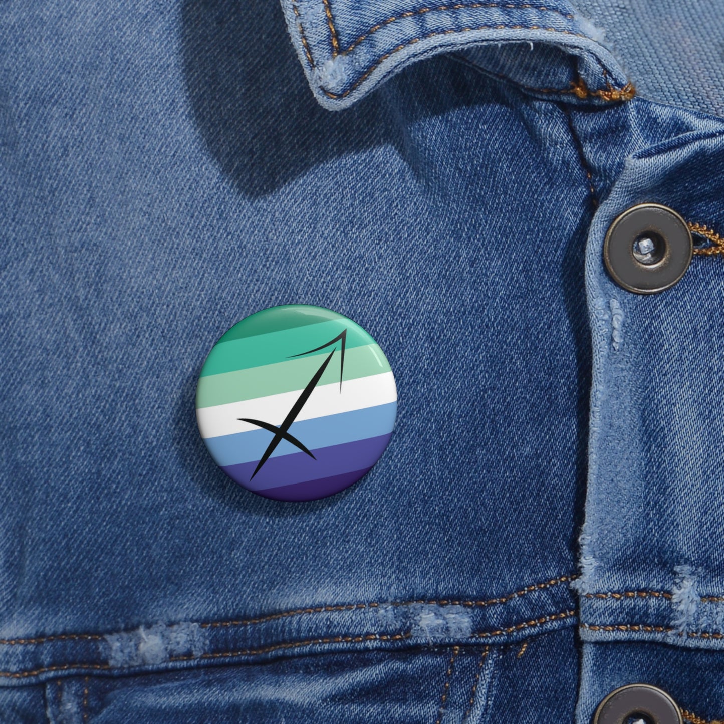 Close-up of a white circular pride button featuring the Sagittarius zodiac glyph centered over a gay men (MLM) pride flag background in greens/teals, white, and blues, attached to a blue denim jacket at the right chest with the button clearly visible.