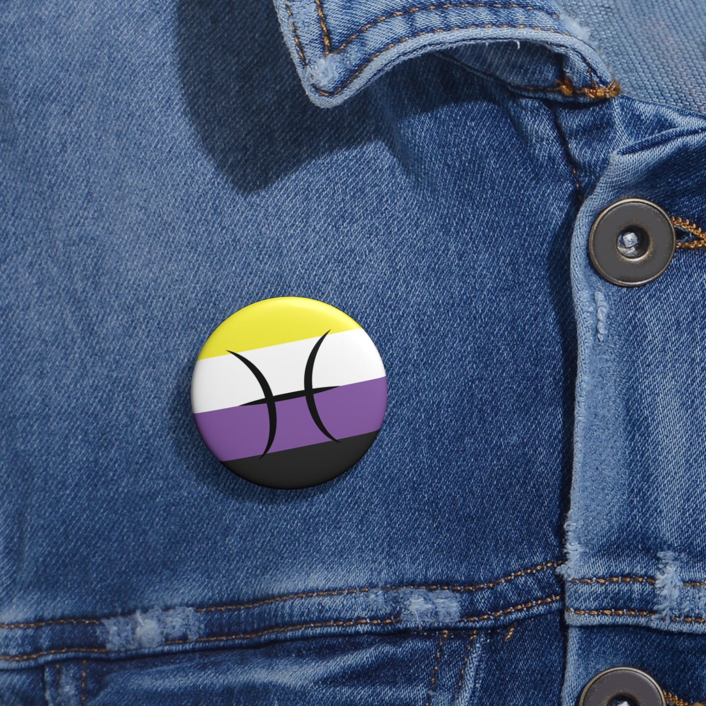 Close-up of a white circular pride button featuring the Pisces zodiac glyph centered over a nonbinary pride flag background in yellow, white, purple, and black, attached to a blue denim jacket at the right chest with the button clearly visible.