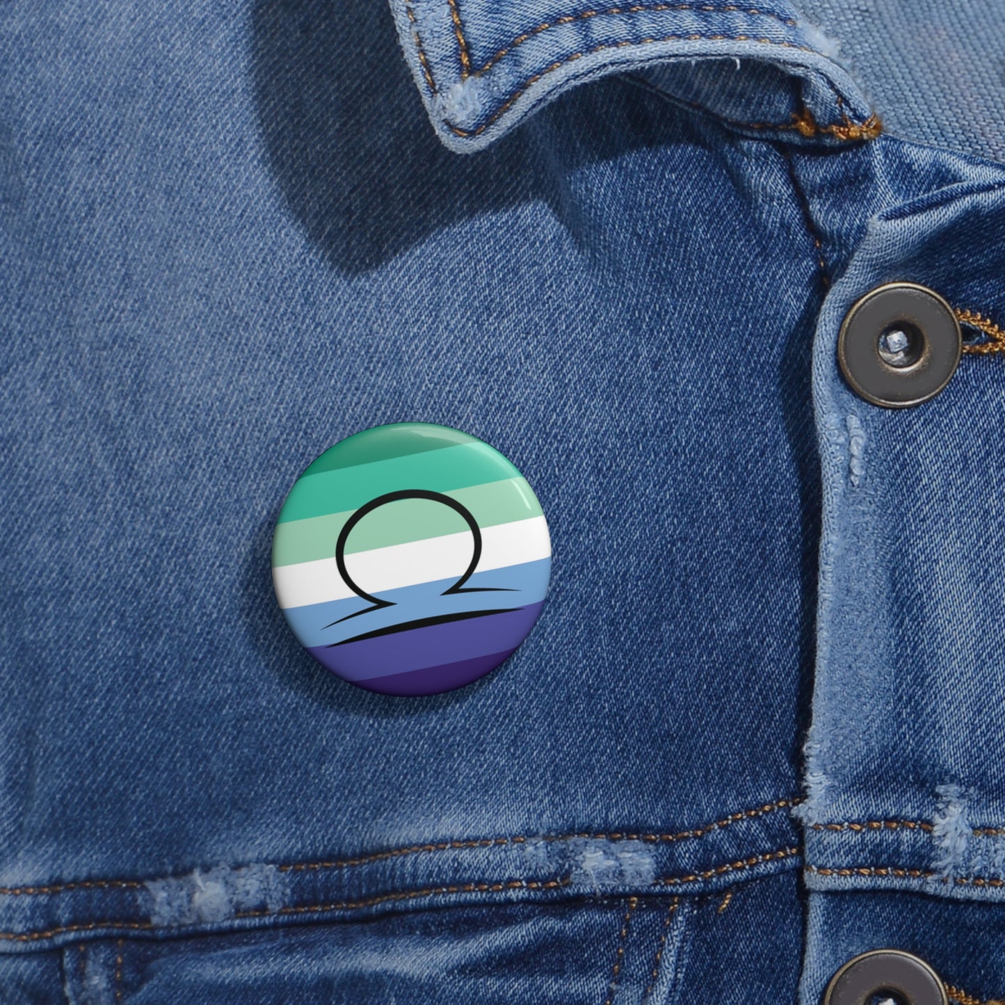 Close-up of a white circular pride button featuring the Libra zodiac glyph centered over a gay men (MLM) pride flag background in greens/teals, white, and blues, attached to a blue denim jacket at the right chest with the button clearly visible.