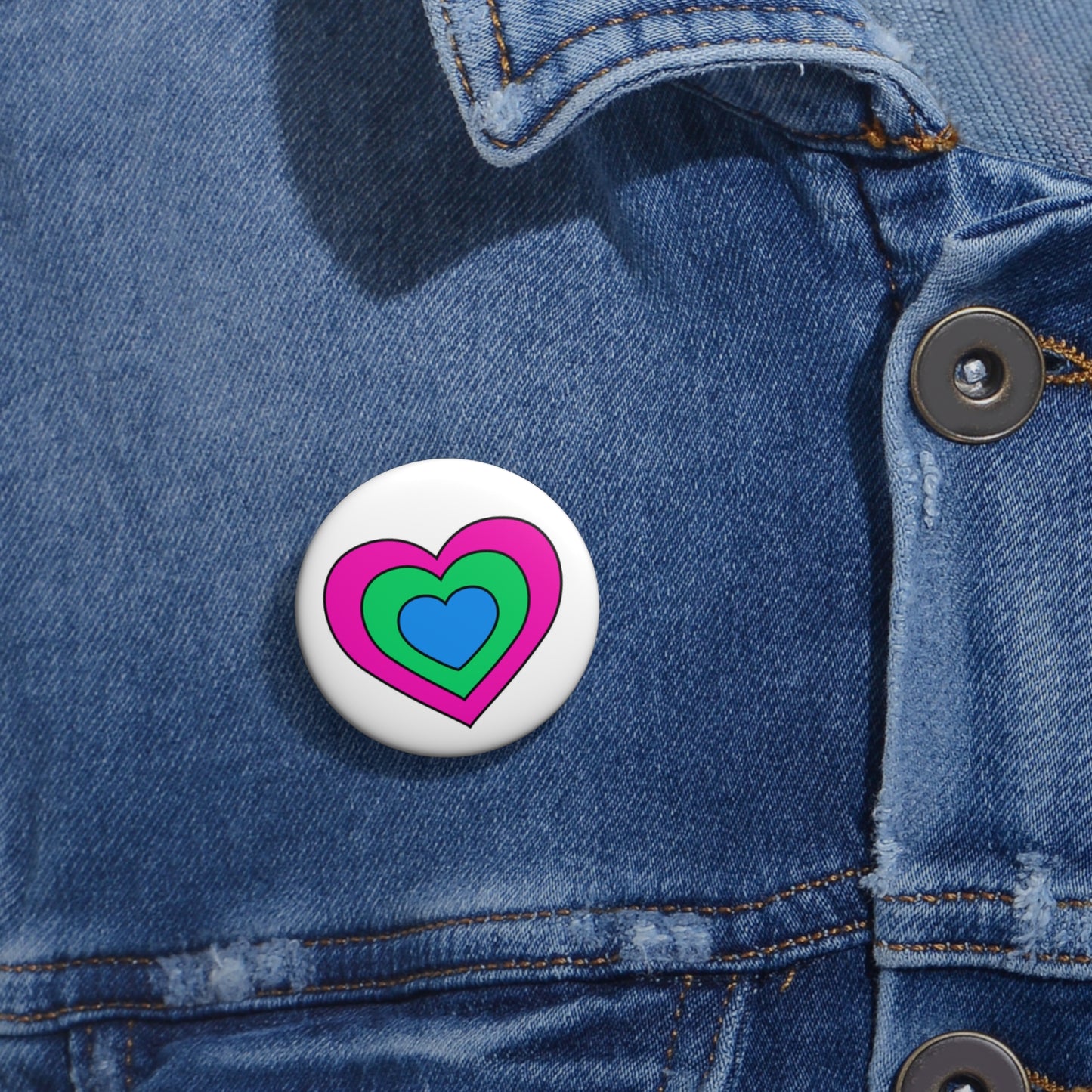 Close-up of a white circular pride button featuring a nested heart design inspired by the Polysexual pride flag in pink, green and blue, attached to a blue denim jacket at the right chest with the button clearly visible.
