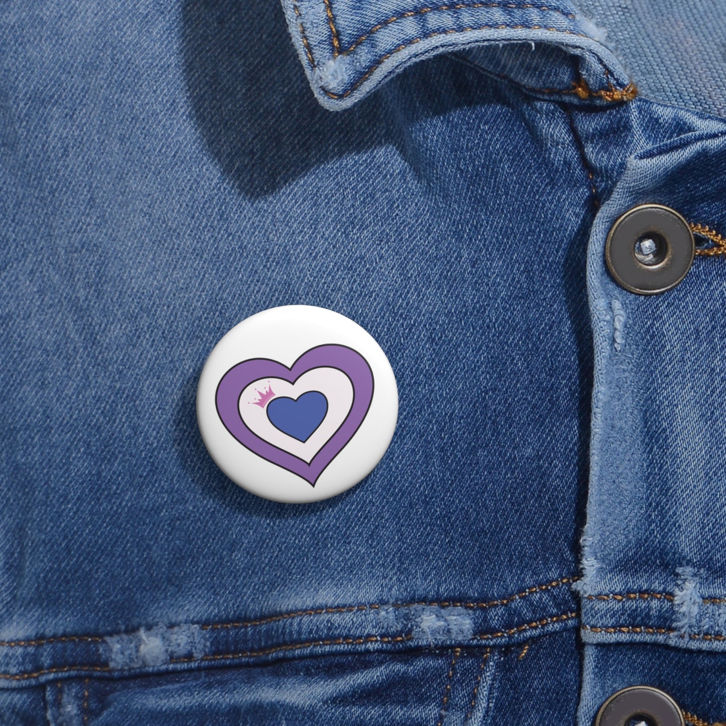 Close-up of a white circular pride button featuring a nested heart design inspired by the Drag pride flag in lavender, white, and blue with a pink crown, attached to a blue denim jacket at the right chest with the button clearly visible.