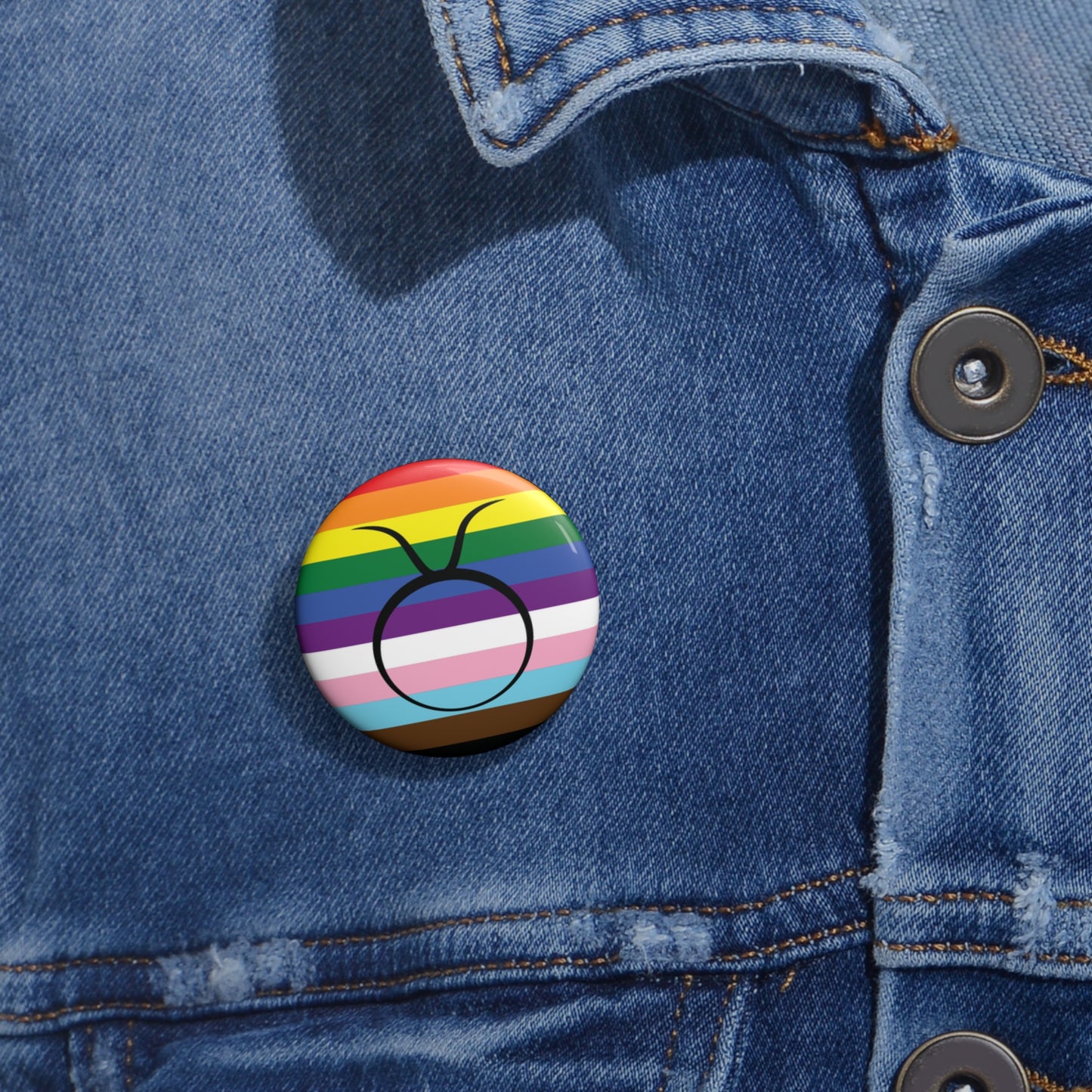 Close-up of a white circular pride button featuring the Taurus zodiac glyph centered over a Progress Pride flag background in rainbow stripes with black, brown, light blue, pink, and white elements, attached to a blue denim jacket at the right chest with the button clearly visible.