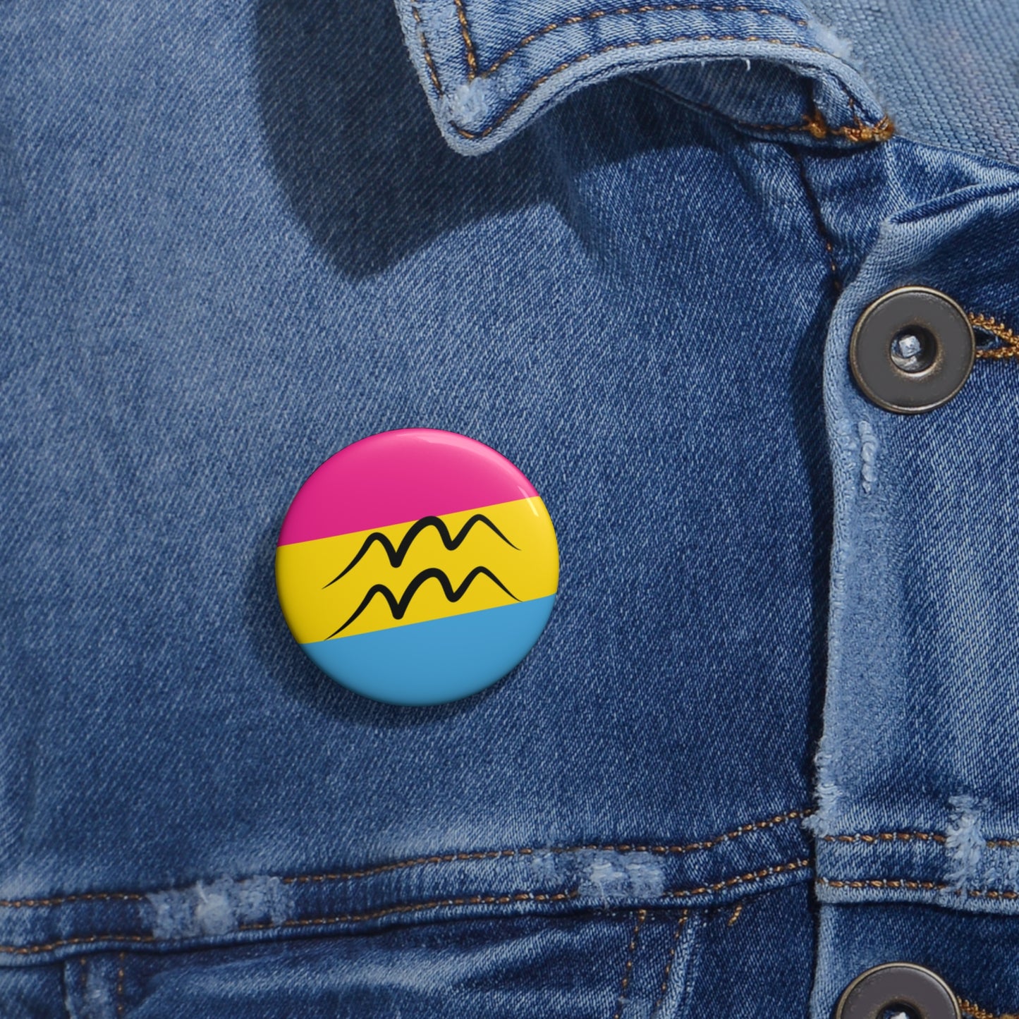 Close-up of a white circular pride button featuring the Aquarius zodiac glyph centered over a Pansexual pride flag background in pink, yellow, and blue, attached to a blue denim jacket at the right chest with the button clearly visible.