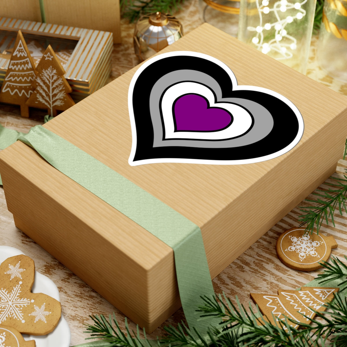 Gift box with a green ribbon on a festive background with cookies and Christmas decorations. Heart-shaped Asexual pride sticker centered on the box.