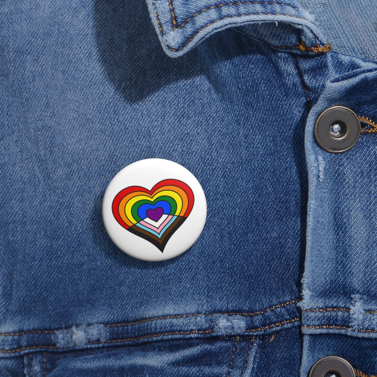 Close-up of a white circular pride button featuring a nested heart design inspired by the Progress pride flag in rainbow, black, brown and trans flag colors, attached to a blue denim jacket at the right chest with the button clearly visible.
