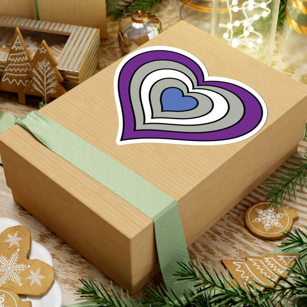 Gift box with a green ribbon on a festive background with cookies and Christmas decorations. Heart-shaped Greysexual pride sticker centered on the box.
