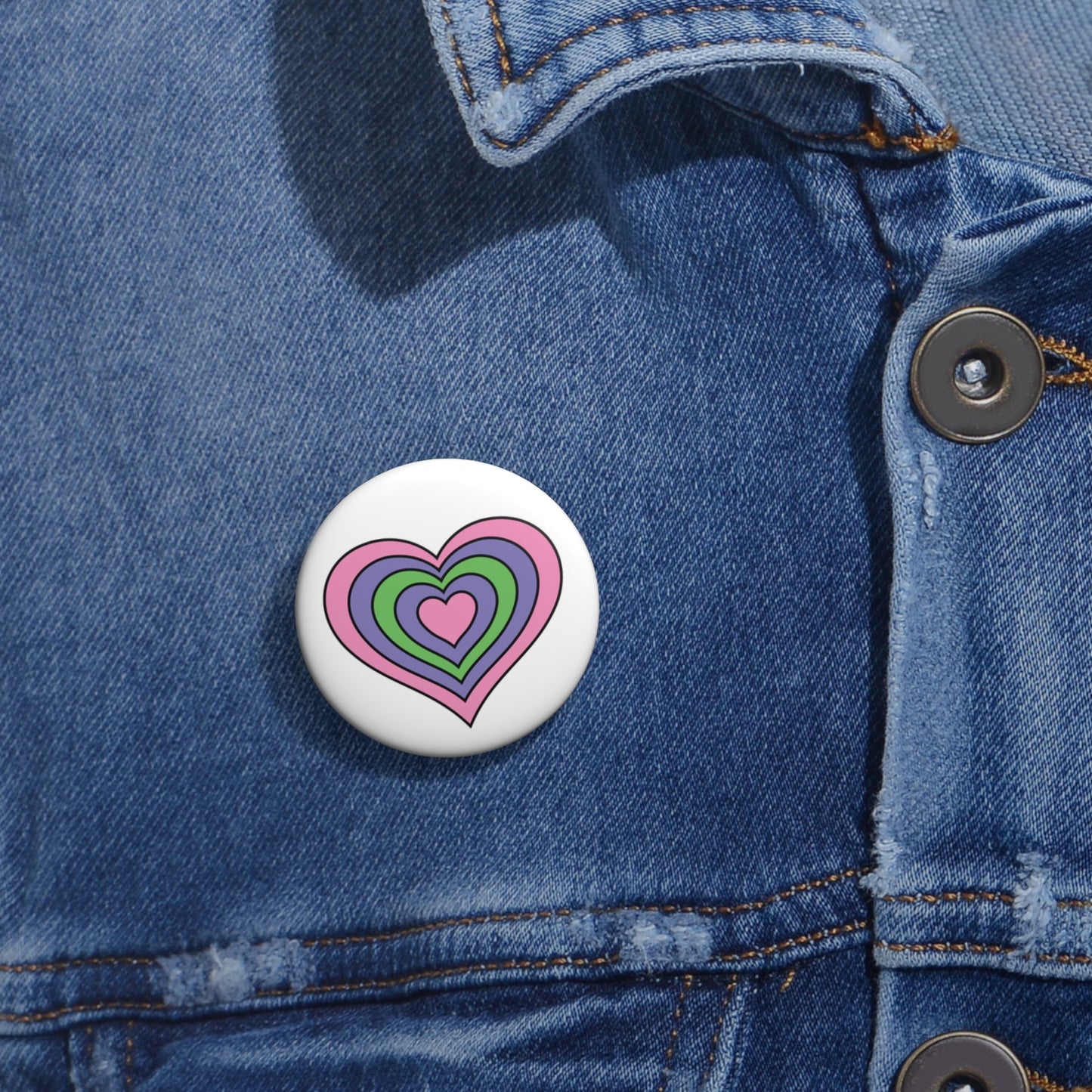 Close-up of a white circular pride button featuring a nested heart design inspired by the Trigender pride flag in pink, purple and green, attached to a blue denim jacket at the right chest with the button clearly visible.