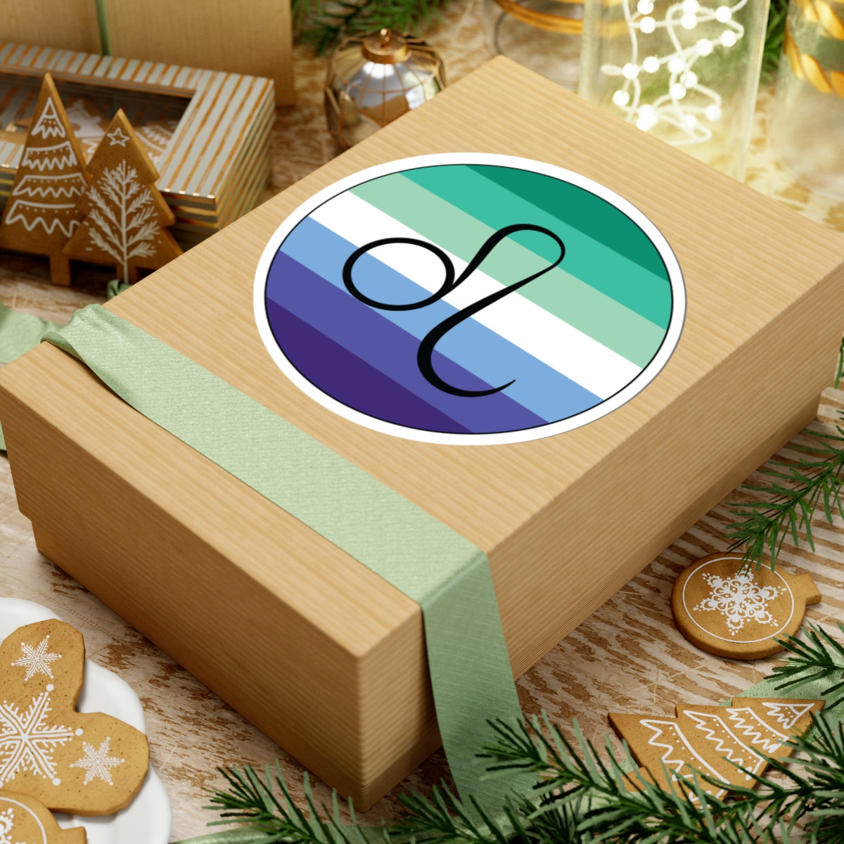 Gift box with a green ribbon on a festive background with cookies and Christmas decorations. Leo Gay Men pride sticker centered on the box.