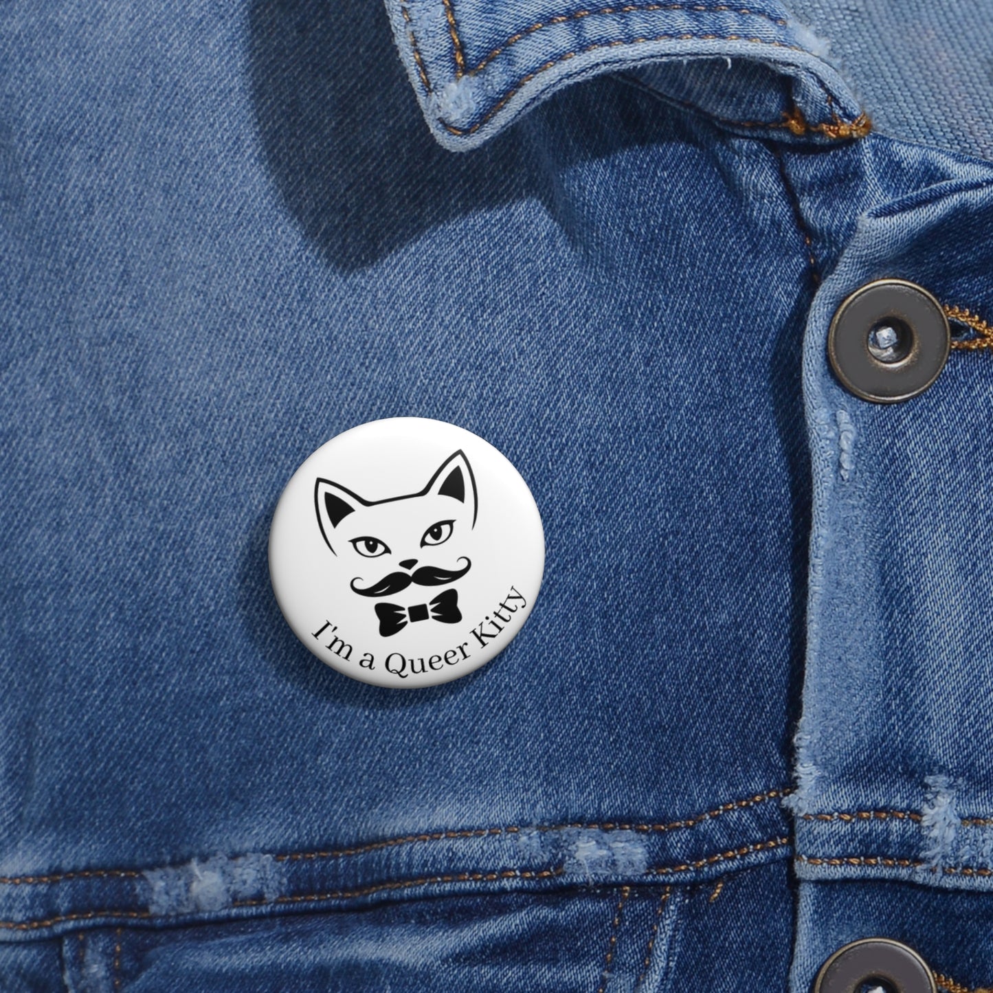 Close-up of a white circular button featuring a the queer kitty logo (a cat face with a mustache and bowtie) with the words, "I'm a Queer Kitty" printed below, attached to a blue denim jacket at the right chest with the button clearly visible.