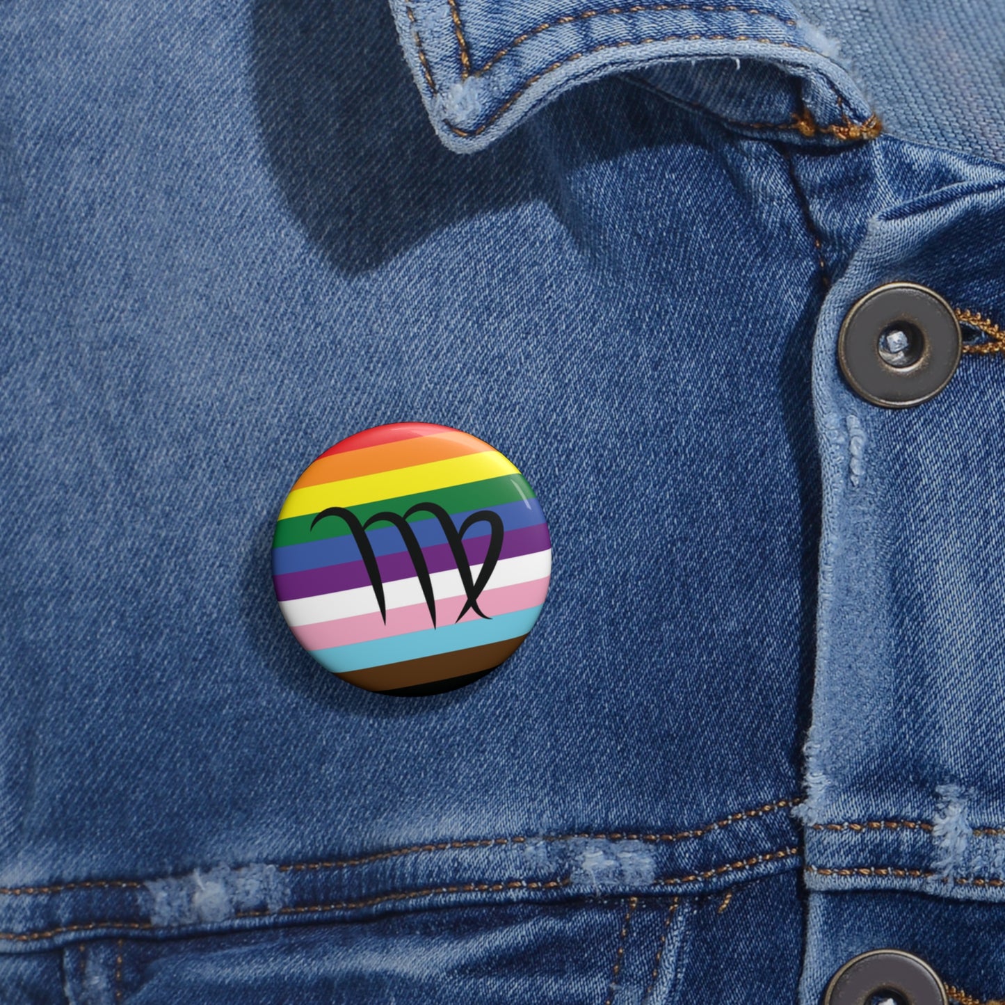 Close-up of a white circular pride button featuring the Virgo zodiac glyph centered over a Progress Pride flag background in rainbow stripes with black, brown, light blue, pink, and white elements, attached to a blue denim jacket at the right chest with the button clearly visible.