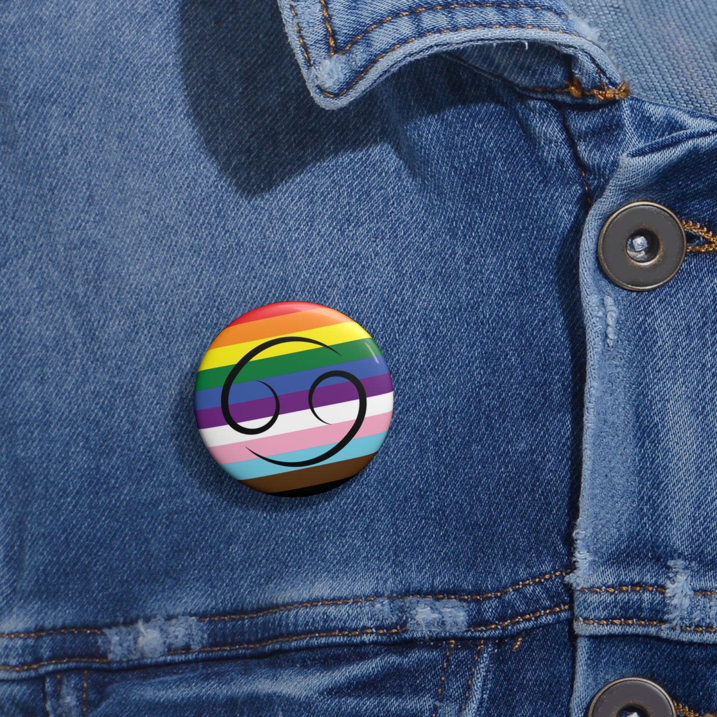 Close-up of a white circular pride button featuring the Cancer zodiac glyph centered over a Progress Pride flag background in rainbow stripes with black, brown, light blue, pink, and white elements, attached to a blue denim jacket at the right chest with the button clearly visible.