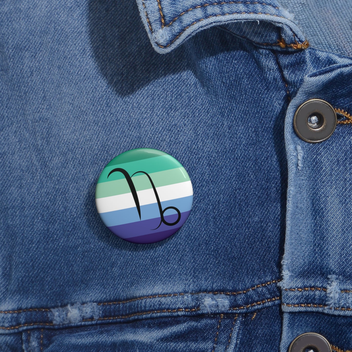 Close-up of a white circular pride button featuring the Capricorn zodiac glyph centered over a gay men (MLM) pride flag background in greens/teals, white, and blues, attached to a blue denim jacket at the right chest with the button clearly visible.