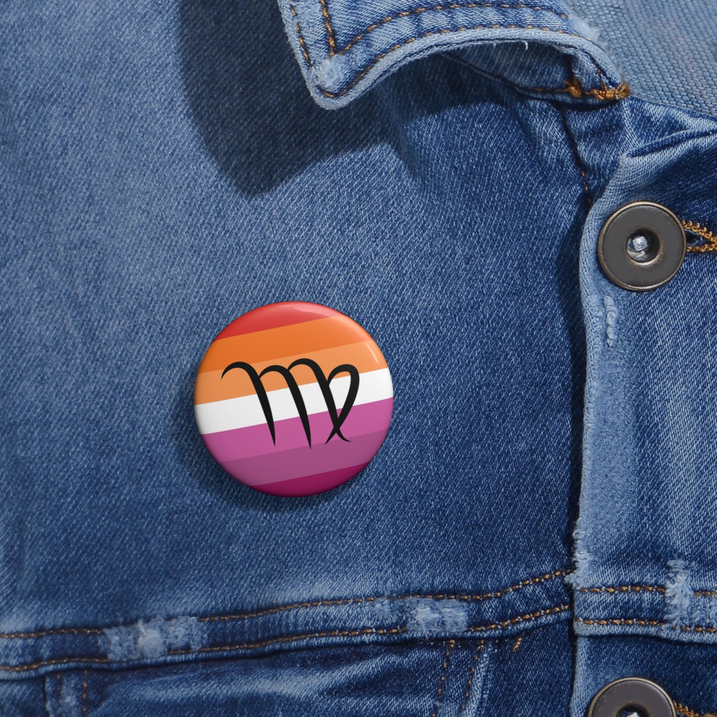 Close-up of a white circular pride button featuring the Virgo zodiac glyph centered over a lesbian pride flag background in oranges, white, and pinks attached to a blue denim jacket at the right chest with the button clearly visible.