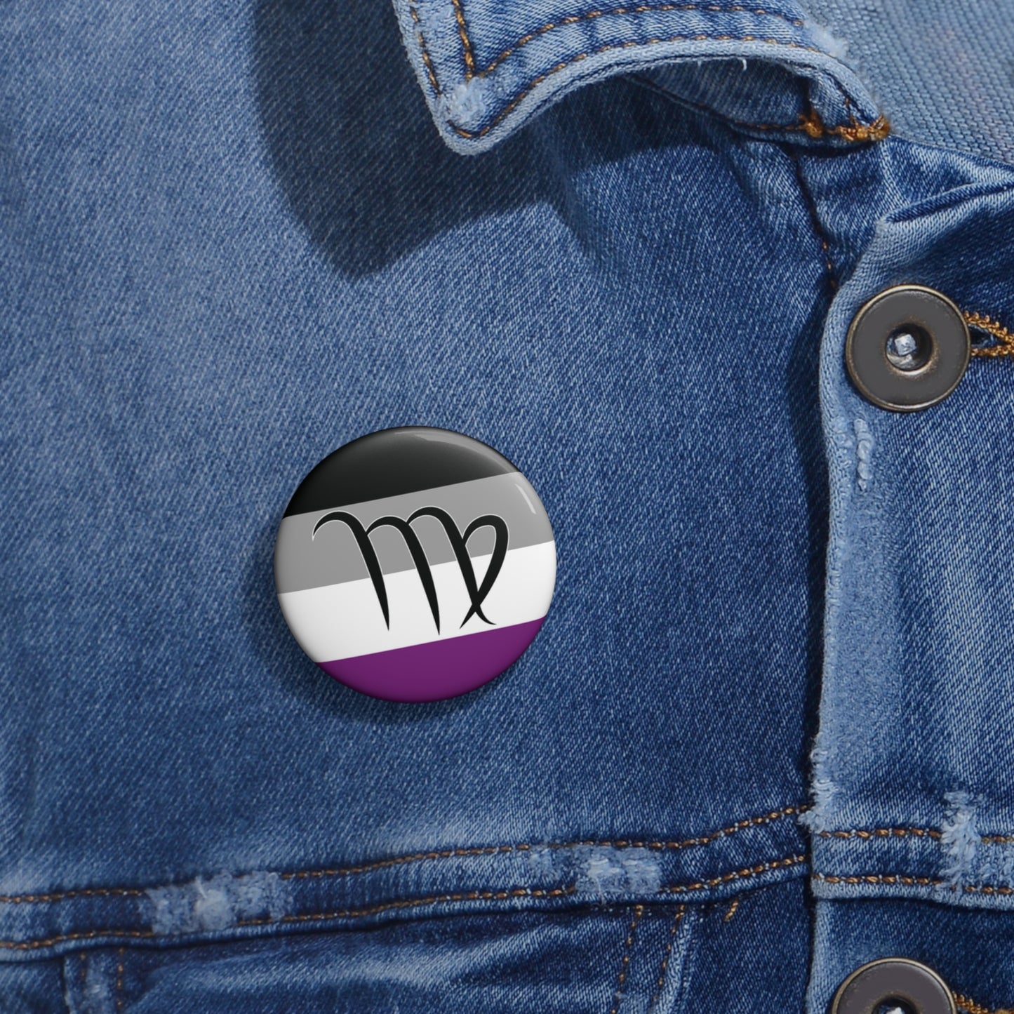 Close-up of a white circular pride button featuring the Virgo zodiac glyph centered over an asexual pride flag background in black, gray, white, and purple, attached to a blue denim jacket at the right chest with the button clearly visible.