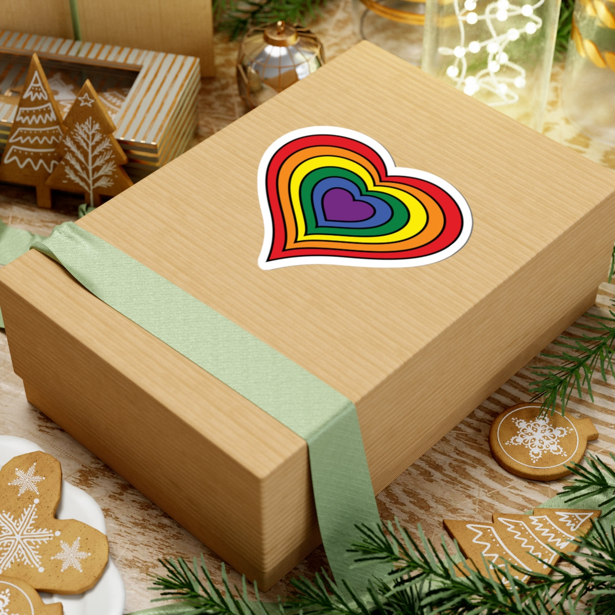 Gift box with a green ribbon on a festive background with cookies and Christmas decorations. Rainbow pride heart sticker centered on the box.