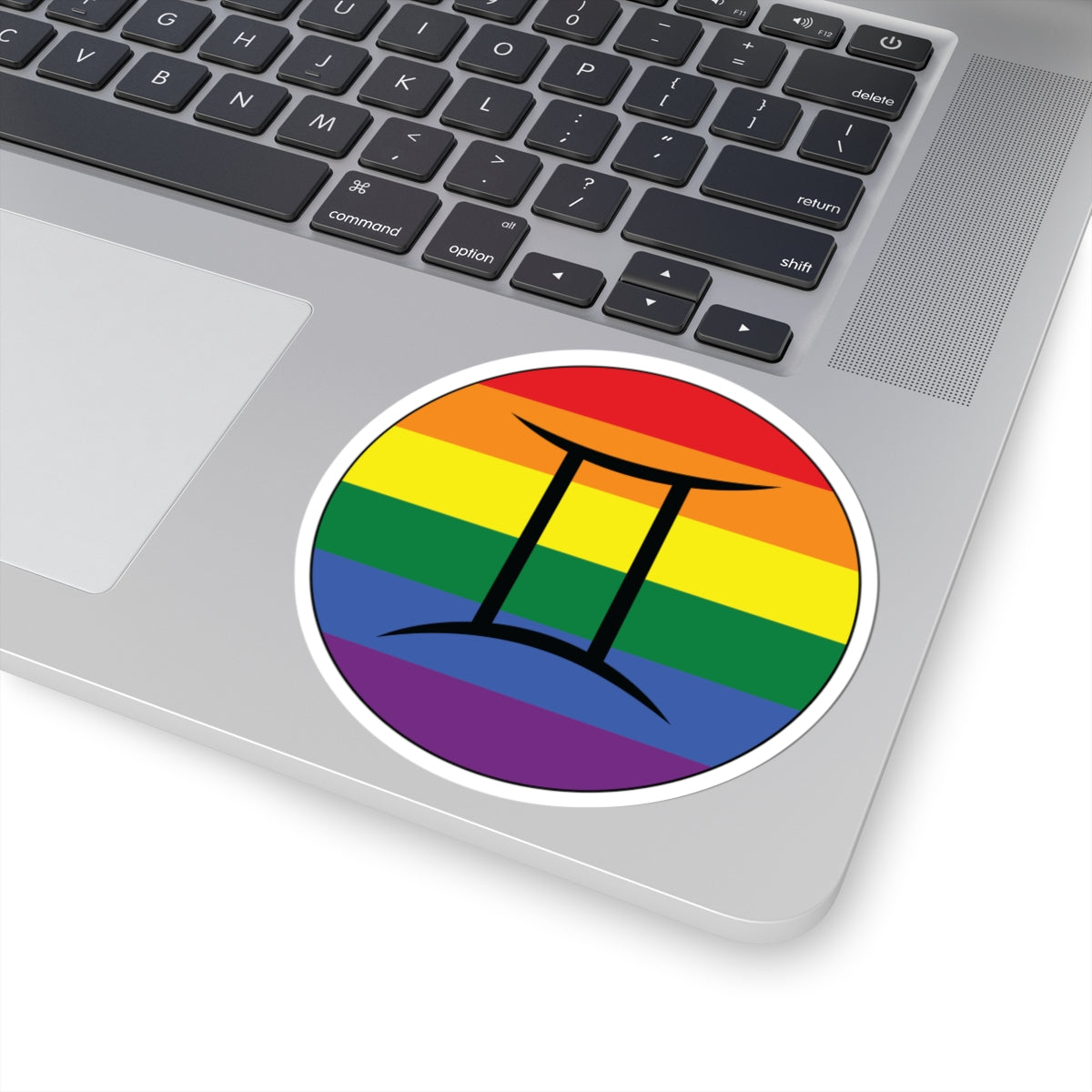 Close-up of the bottom right corner of a silver laptop featuring a 4 inch by 4 inch circular zodiac sticker with a centered Gemini glyph on a Rainbow pride flag style background applied to the surface.