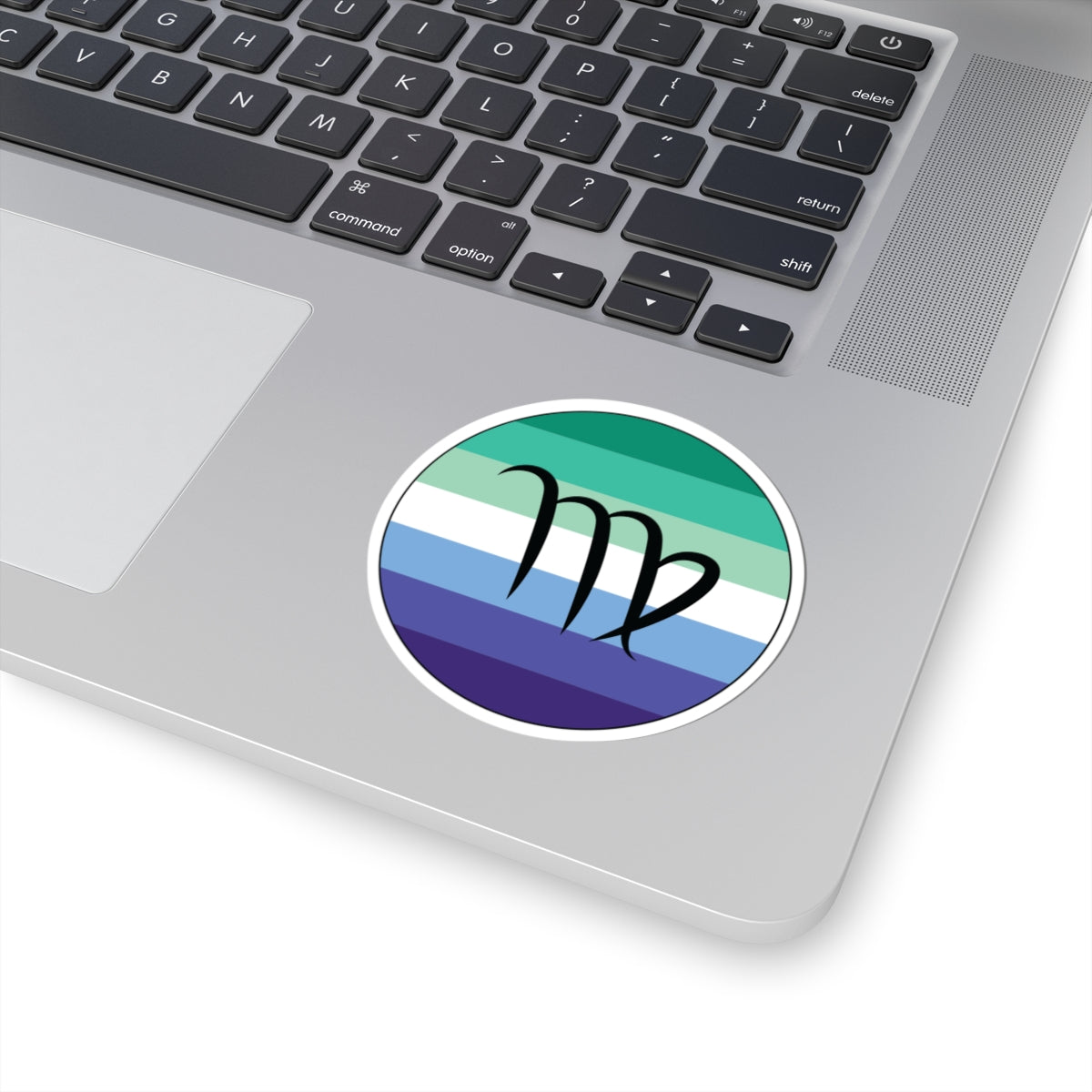 Close-up of the bottom right corner of a silver laptop featuring a 3 inch by 3 inch circular zodiac sticker with a centered Virgo glyph on a Gay Men pride flag style background applied to the surface.
