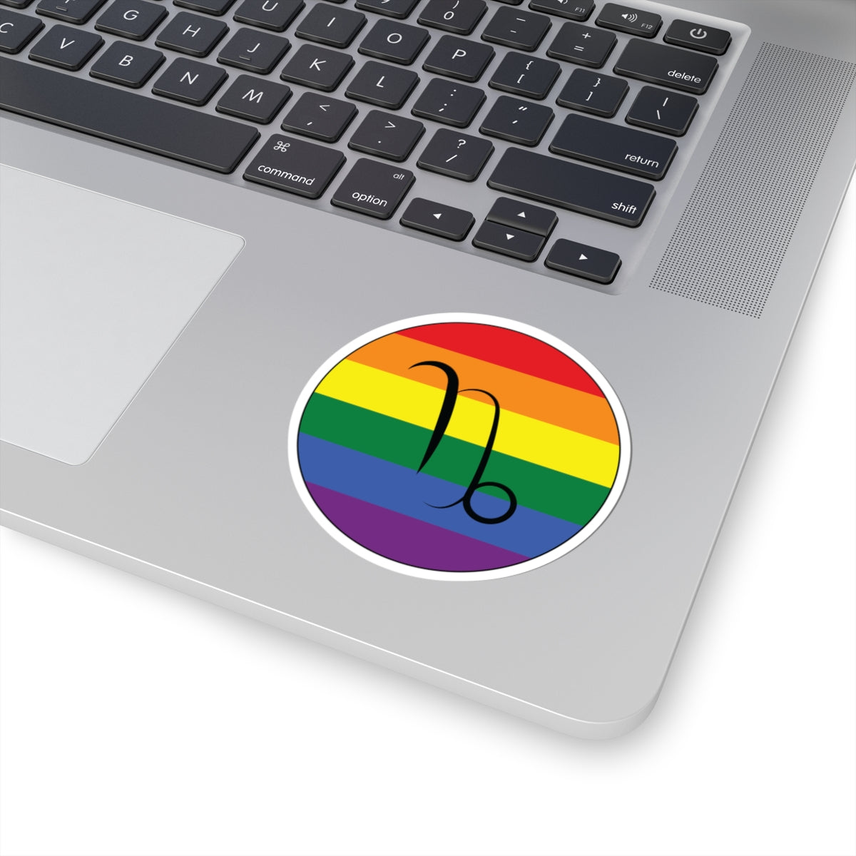 Close-up of the bottom right corner of a silver laptop featuring a 3 inch by 3 inch circular zodiac sticker with a centered Capricorn glyph on a Rainbow pride flag style background applied to the surface.