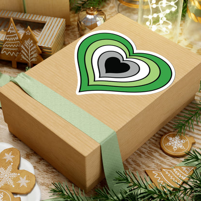 Gift box with a green ribbon on a festive background with cookies and Christmas decorations. Heart-shaped Aromantic pride sticker centered on the box.