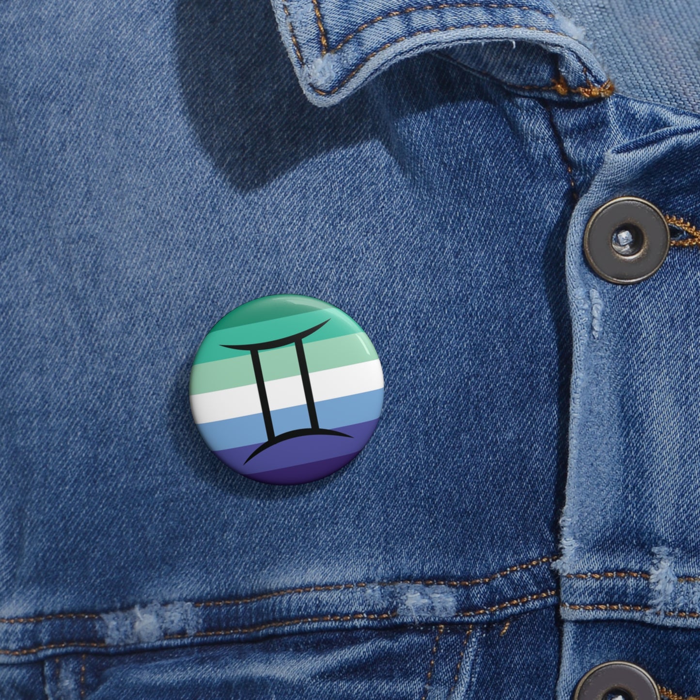 Close-up of a white circular pride button featuring the Gemini zodiac glyph centered over a gay men (MLM) pride flag background in greens/teals, white, and blues, attached to a blue denim jacket at the right chest with the button clearly visible.