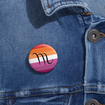 Close-up of a white circular pride button featuring the Scorpio zodiac glyph centered over a lesbian pride flag background in oranges, white, and pinks attached to a blue denim jacket at the right chest with the button clearly visible.