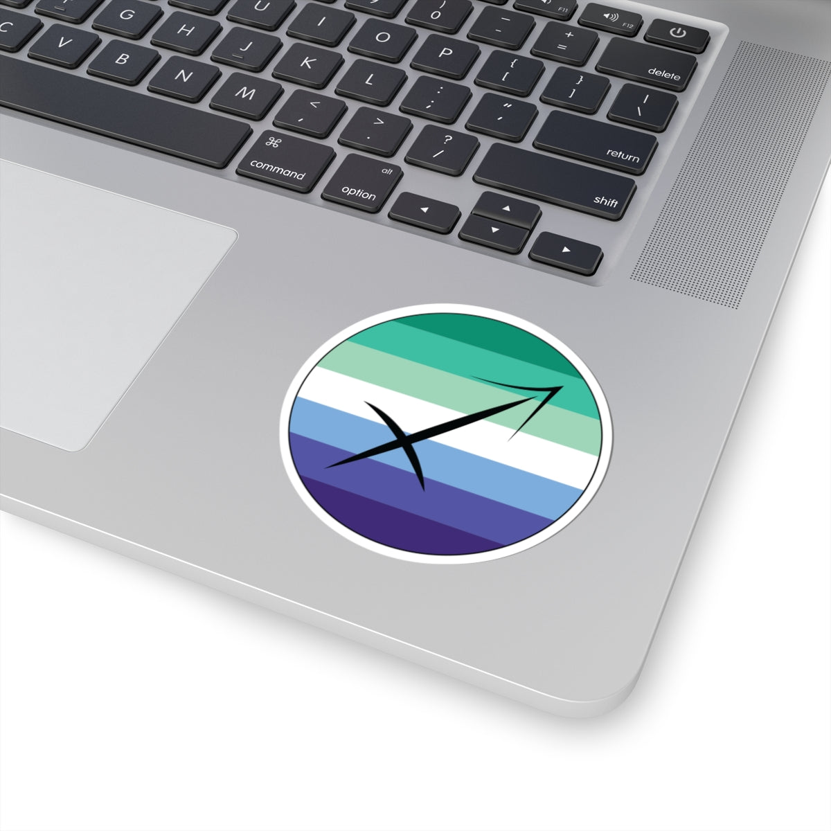 Close-up of the bottom right corner of a silver laptop featuring a 3 inch by 3 inch circular zodiac sticker with a centered Sagittarius glyph on a Gay Men pride flag style background applied to the surface.