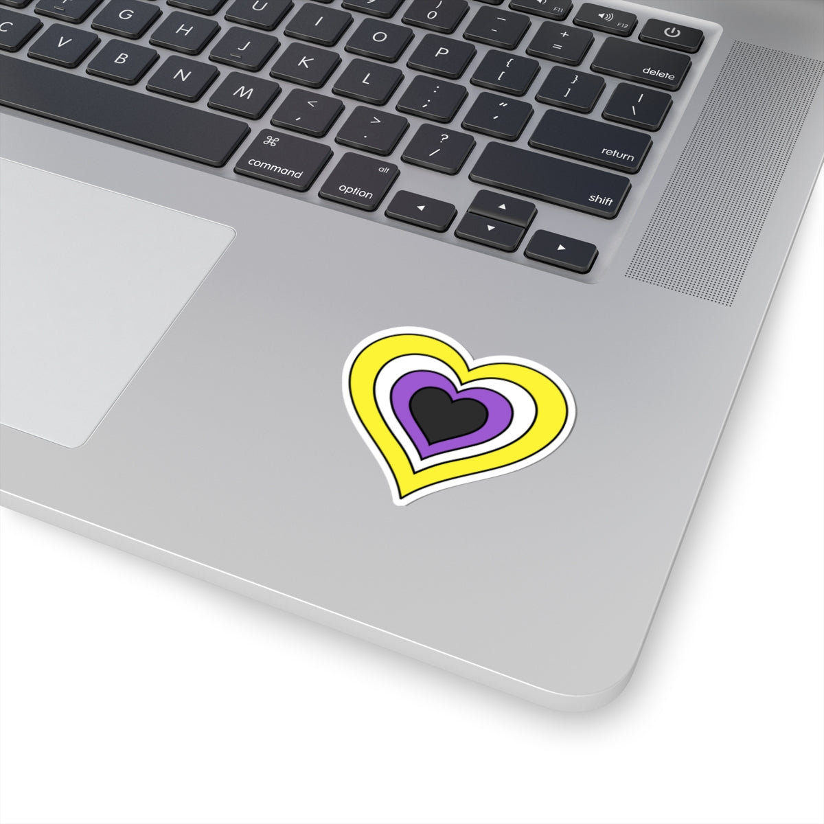 Close-up of the bottom right corner of a silver laptop featuring a 2 inch by 2 inch pride flag heart sticker in a Nonbinary flag style applied to the surface.