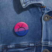 Close-up of a white circular pride button featuring the Taurus zodiac glyph centered over a bisexual pride flag background in pink, purple, and blue, attached to a blue denim jacket at the right chest with the button clearly visible.