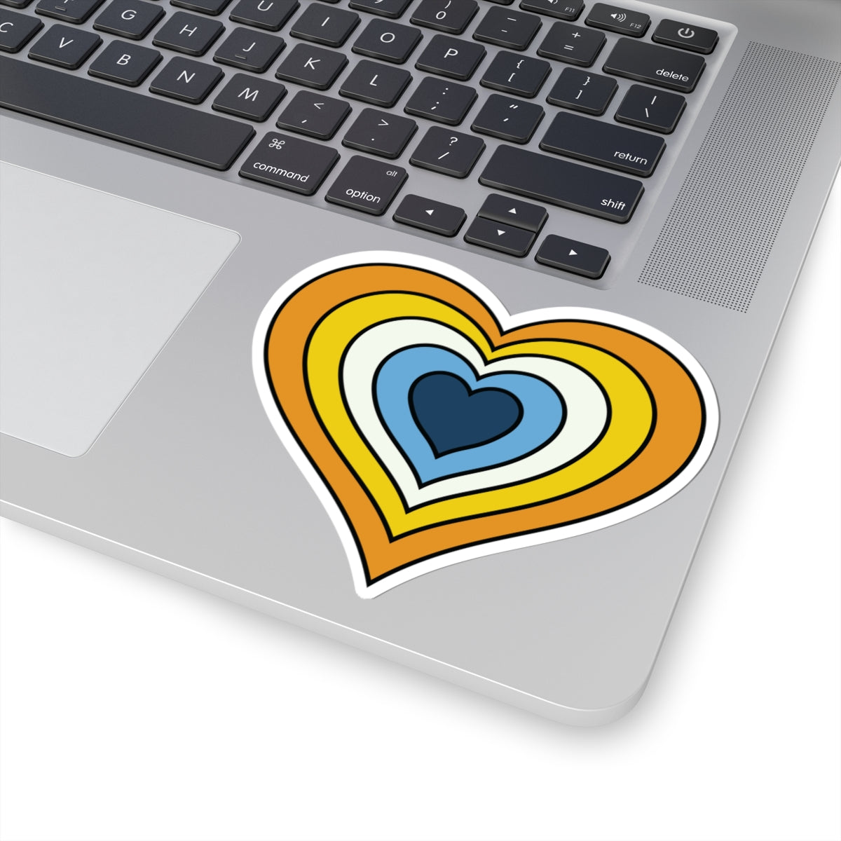 Close-up of the bottom right corner of a silver laptop featuring a 4 inch by 4 inch pride flag heart sticker in an Aroace flag style applied to the surface.