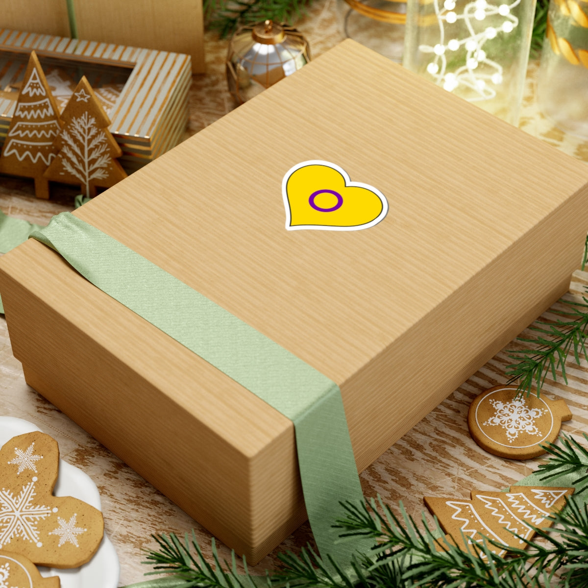 Gift box with a green ribbon on a festive background with cookies and Christmas decorations. Heart-shaped Intersex pride sticker centered on the box.