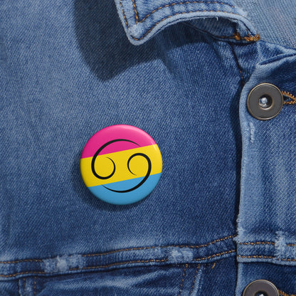 Close-up of a white circular pride button featuring the Cancer zodiac glyph centered over a Pansexual pride flag background in pink, yellow, and blue, attached to a blue denim jacket at the right chest with the button clearly visible.