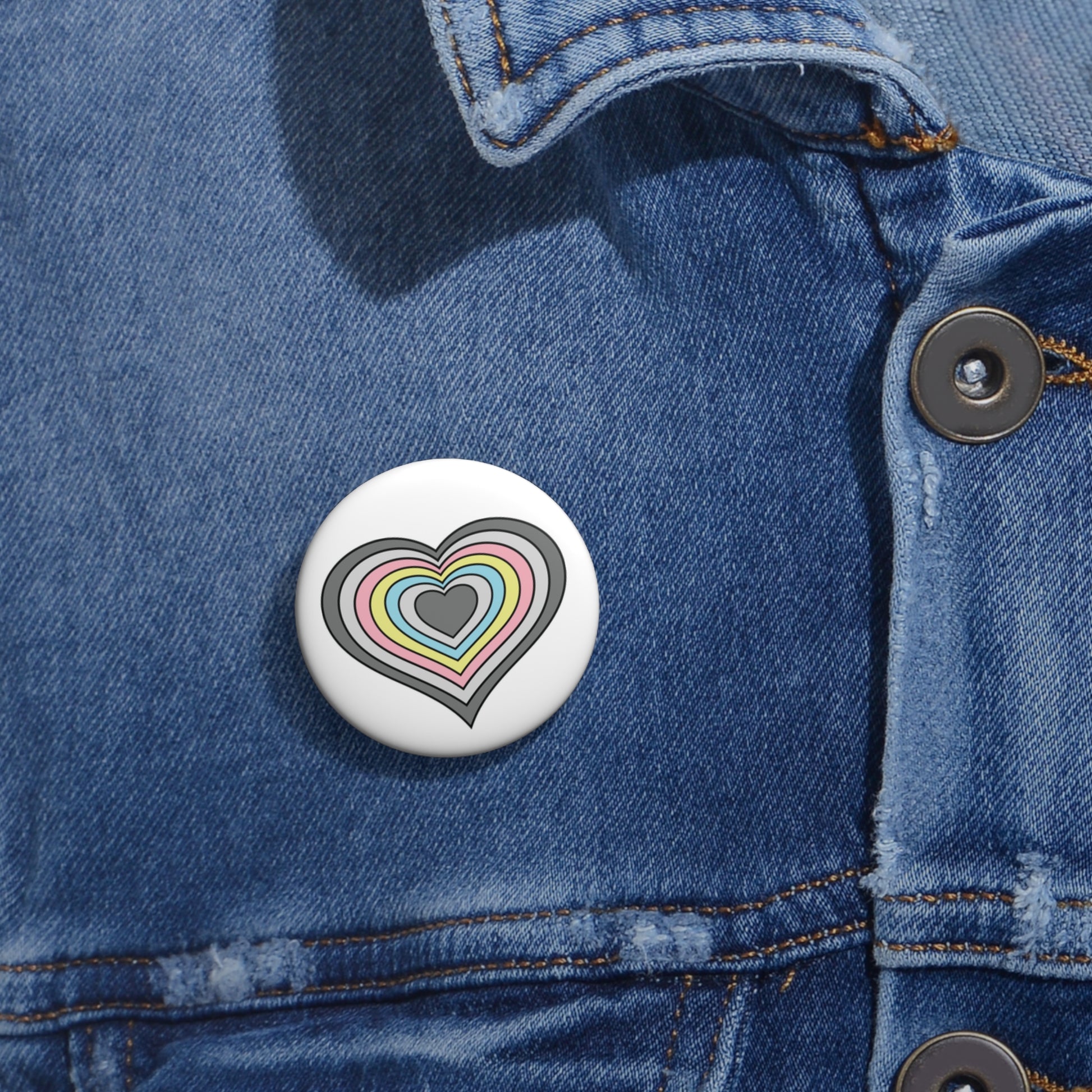 Close-up of a white circular pride button featuring a nested heart design inspired by the Demiflux pride flag in grays, pink, yellow and blue, attached to a blue denim jacket at the right chest with the button clearly visible.