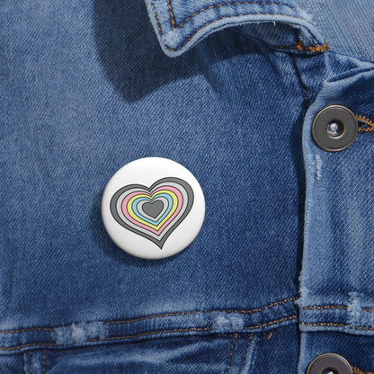 Close-up of a white circular pride button featuring a nested heart design inspired by the Demiflux pride flag in grays, pink, yellow and blue, attached to a blue denim jacket at the right chest with the button clearly visible.