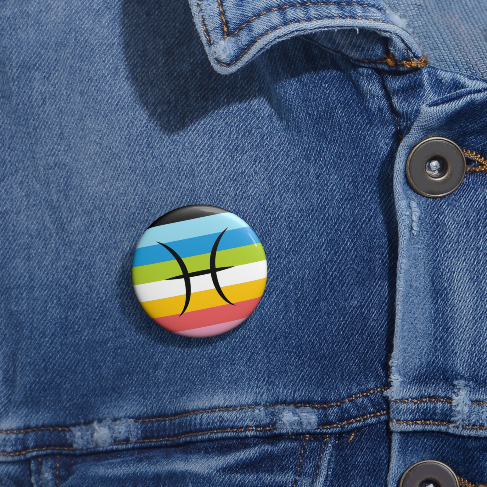 Close-up of a white circular pride button featuring the Pisces zodiac glyph centered over a queer pride flag background in pinks, yellow, white, green, blues, and black, attached to a blue denim jacket at the right chest with the button clearly visible.