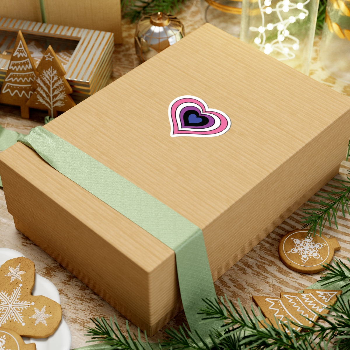 Gift box with a green ribbon on a festive background with cookies and Christmas decorations. Heart-shaped Genderfluid pride sticker centered on the box.