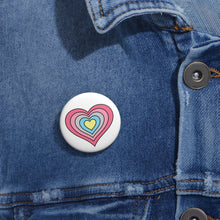 Close-up of a white circular pride button featuring a nested heart design inspired by the Genderflux pride flag in pinks, gray, blue and yellow, attached to a blue denim jacket at the right chest with the button clearly visible.