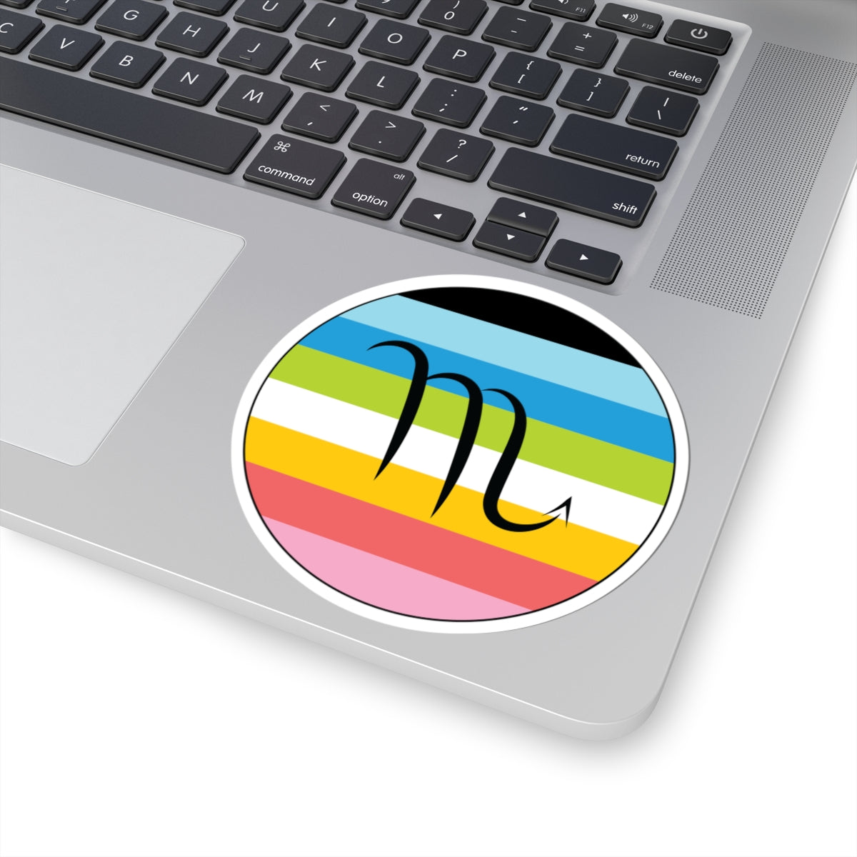 Close-up of the bottom right corner of a silver laptop featuring a 4 inch by 4 inch circular zodiac sticker with a centered Scorpio glyph on a Queer pride flag style background applied to the surface.