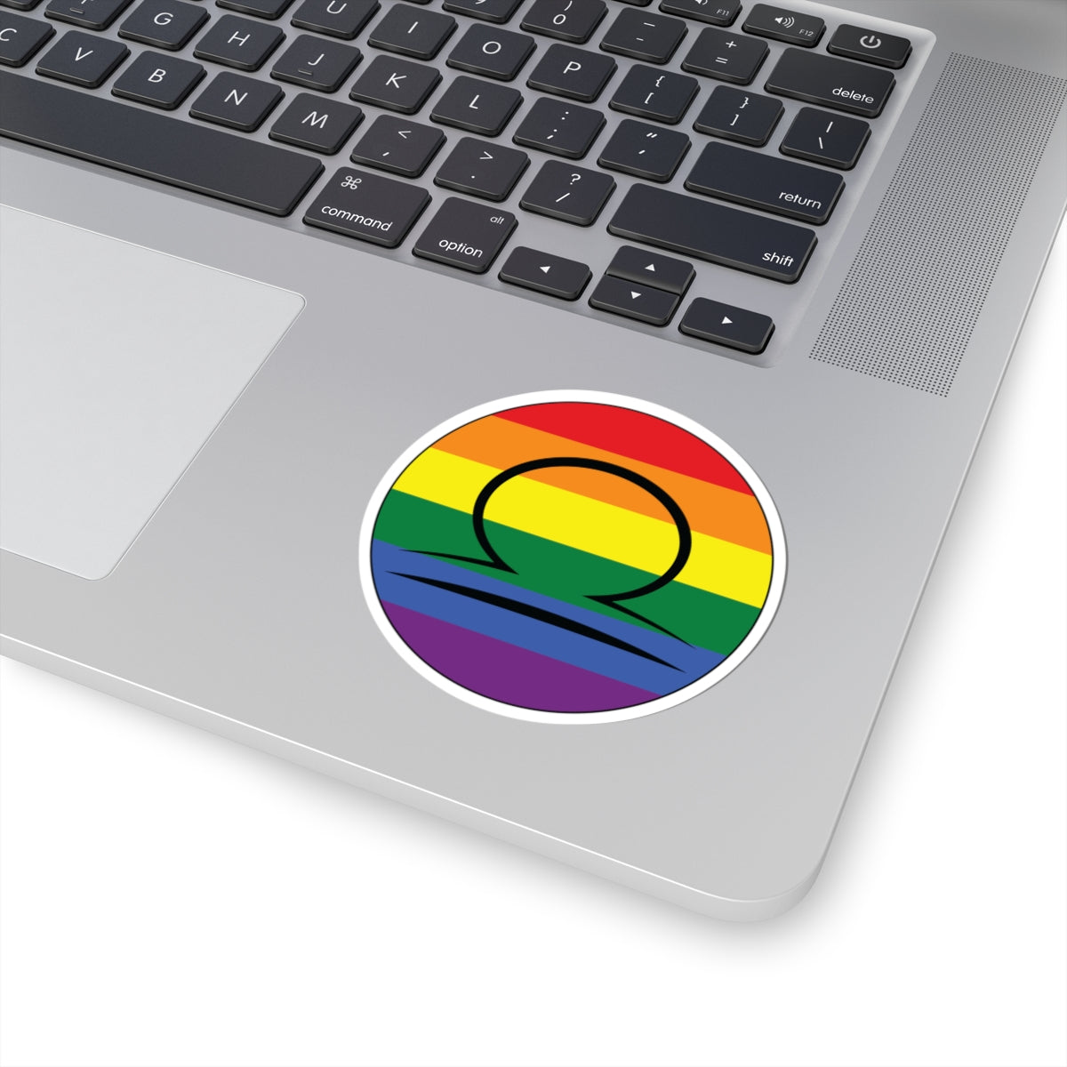 Close-up of the bottom right corner of a silver laptop featuring a 3 inch by 3 inch circular zodiac sticker with a centered Libra glyph on a Rainbow pride flag style background applied to the surface.