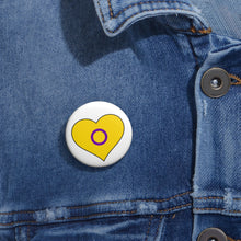 Close-up of a white circular pride button featuring a nested heart design inspired by the Intersex pride flag in yellow and purple, attached to a blue denim jacket at the right chest with the button clearly visible.