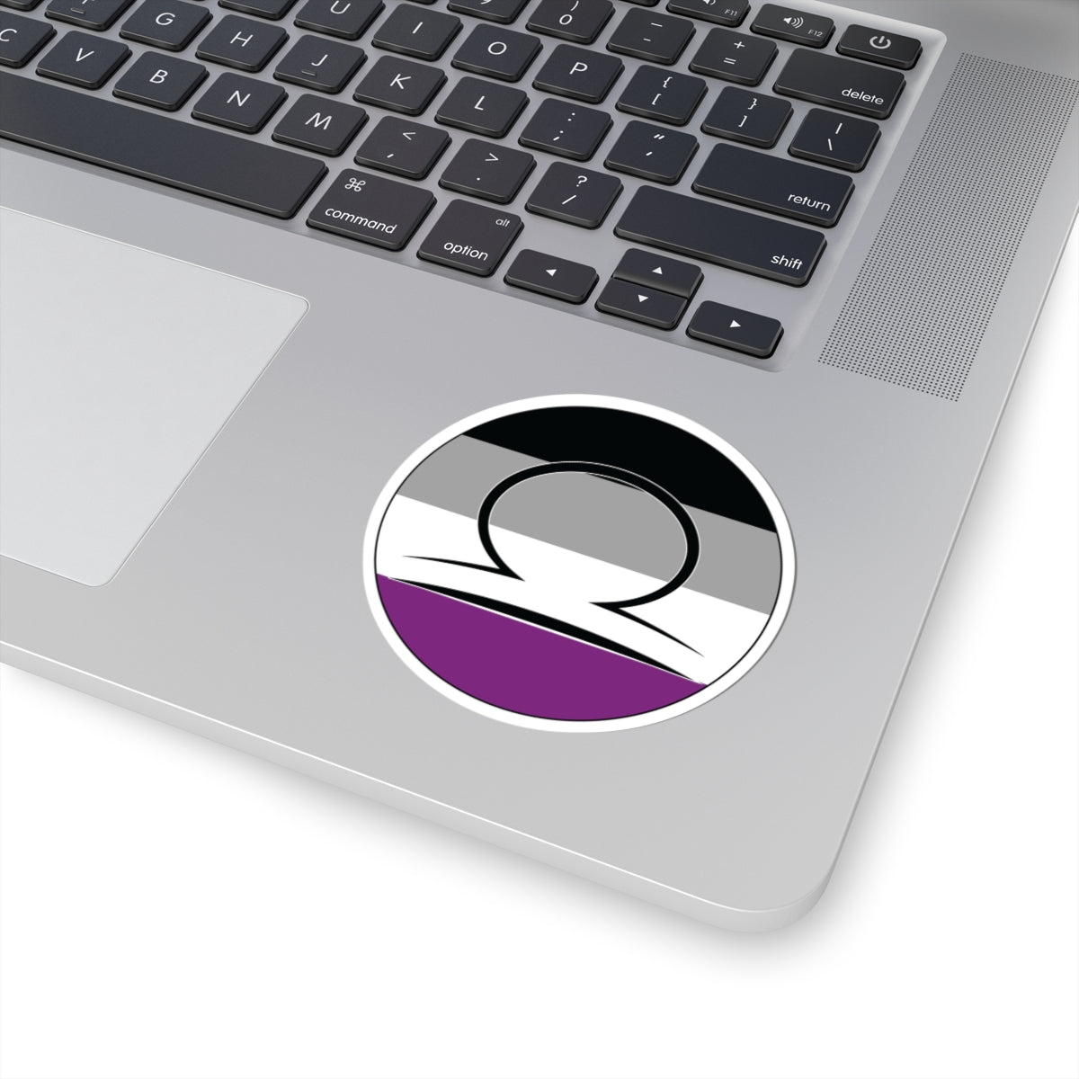 Close-up of the bottom right corner of a silver laptop featuring a 3 inch by 3 inch circular zodiac sticker with a centered Libra glyph on an Asexual pride flag style background applied to the surface.