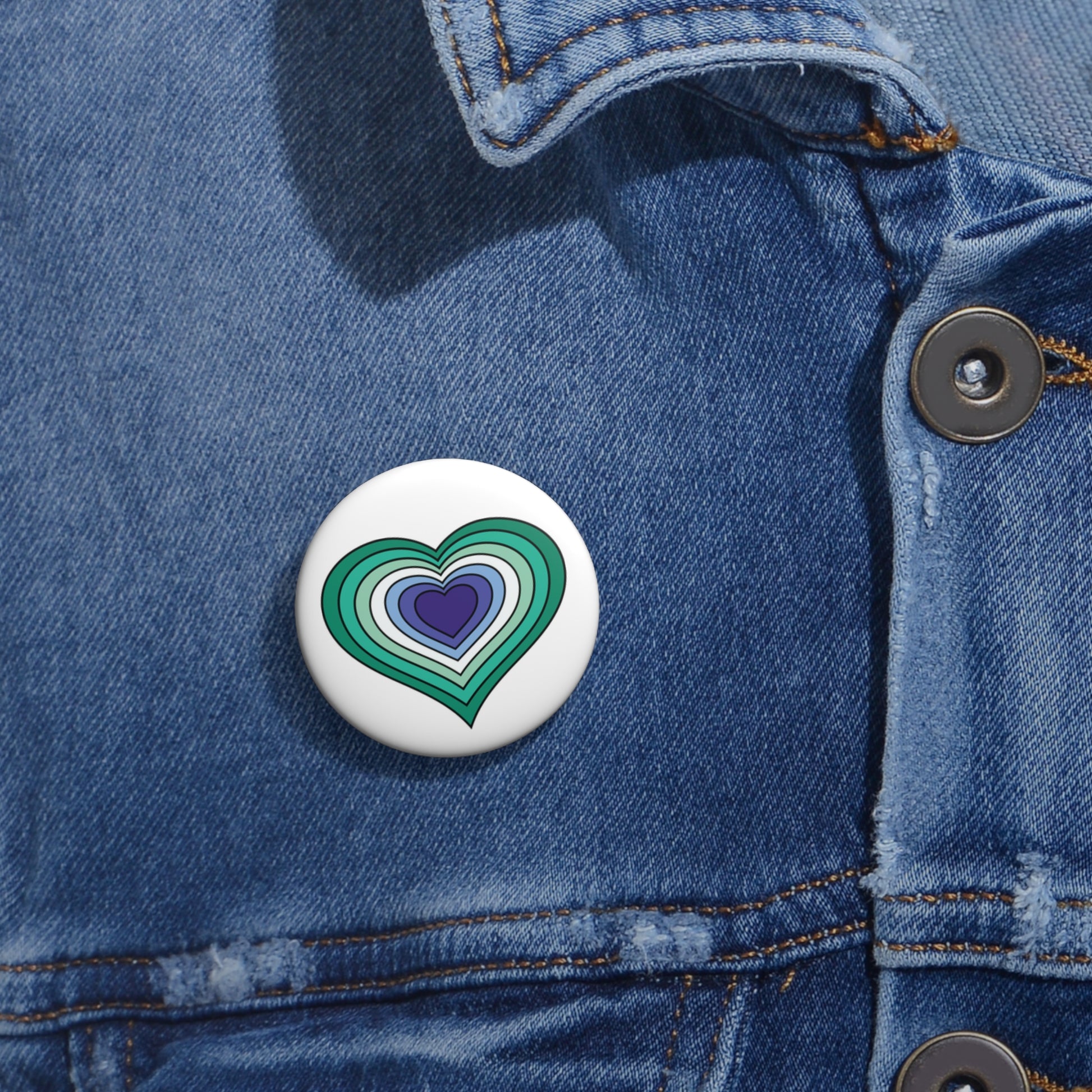 Close-up of a white circular pride button featuring a nested heart design inspired by the Gay Men pride flag in teals/greens, white, and blues, attached to a blue denim jacket at the right chest with the button clearly visible.