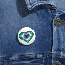 Close-up of a white circular pride button featuring a nested heart design inspired by the Gay Men pride flag in teals/greens, white, and blues, attached to a blue denim jacket at the right chest with the button clearly visible.