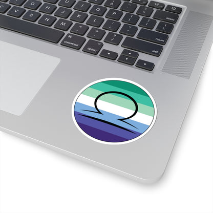Close-up of the bottom right corner of a silver laptop featuring a 3 inch by 3 inch circular zodiac sticker with a centered Libra glyph on a Gay Men pride flag style background applied to the surface.